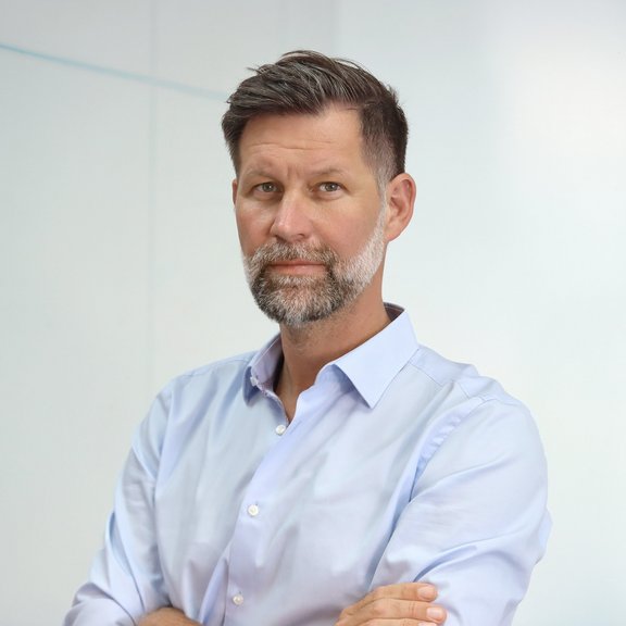 The picture shows a man with brown hair and a beard. He is wearing a light blue shirt and smiling at the camera.