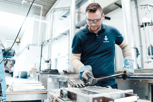 Auf dem Bild ist ein Mitarbeiter in der Produktion beim Gießen von Lotbarren zu sehen. Er trägt ein blaues T-Shirt, Arbeitshandschuhe und eine Schutzbrille.