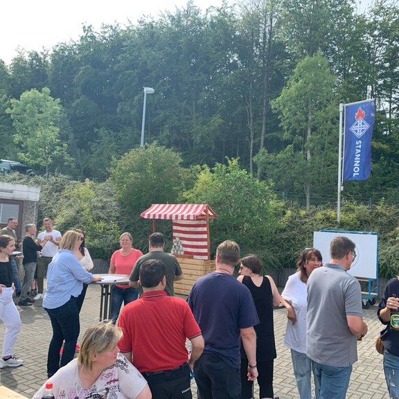 The picture shows several people at a party. A can throwing stand and a target for archery can be seen in the background.