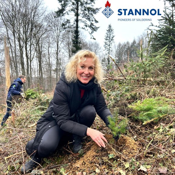 Ein blonde Frau pflanzt einen Baum im Wald. Sie trägt dunkle Kleidung und lächelt freundlich in die Kamera. Im Hintergrund ist eine weitere Person zu sehen, die einen Baum trägt.