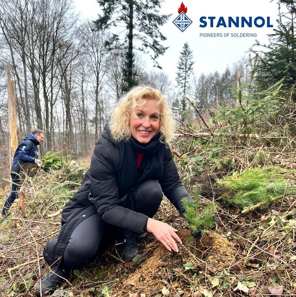 Ein blonde Frau pflanzt einen Baum im Wald. Sie trägt dunkle Kleidung und lächelt freundlich in die Kamera. Im Hintergrund ist eine weitere Person zu sehen, die einen Baum trägt.