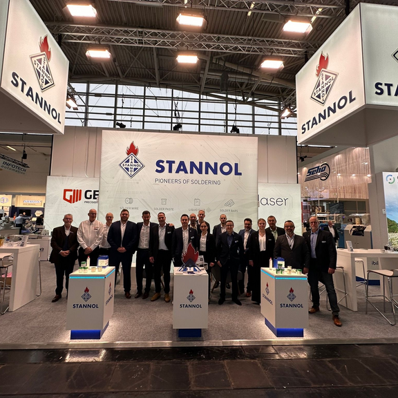 The picture shows the Stannol trade fair stand. In the foreground is a large group of people smiling friendly into the camera.