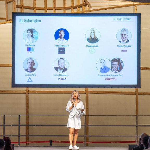 Auf dem Bild ist die Moderatorin der Veranstaltung auf der Bühne zu sehen: Sie ist blond und trägt ein knielanges Kleid. Im Hintergrund ist eine Leinwand mit den Porträts aller Referentinnen und Referenten zu sehen.
