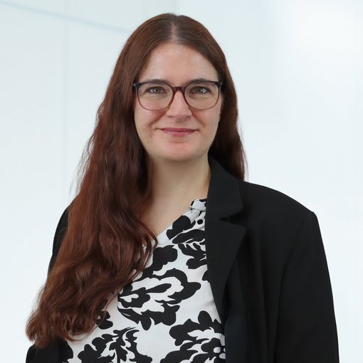 Auf dem Bild ist eine Frau mit langen rotbraunen Haaren und Brille zu sehen Sie trägt eine schwarz-weiß gemusterte Bluse und einen schwarzen Blazer. Sie lächelt freundlich in die Kamera.