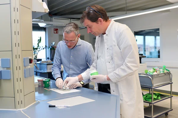 Auf dem Bild sind zwei Männer in einem Labor zu sehen, die mit Lotpaste experimentieren. Beide tragen Einweg-Handschuhe. Der Herr links hat graue Haare, trägt eine Brille und ein hellblaues Hemd. Der Herr rechts trägt ein weißes Shirt und ienen Laborkittel.