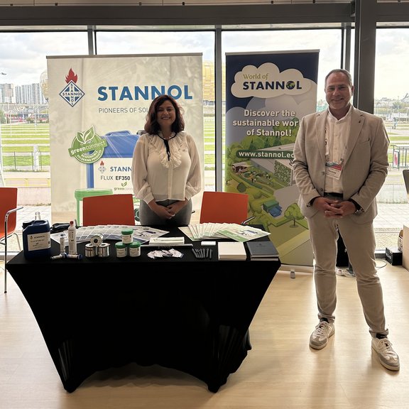 The picture shows a man and a woman in formal attire smiling at the camera. They are standing behind a presentation stand at an event. Stannol solder material is presented on the table. Two Stannol display stands can be seen in the background.