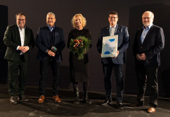 The picture shows five people against a dark background. Three of them are holding a certificate, a trophy and a bouquet of flowers.