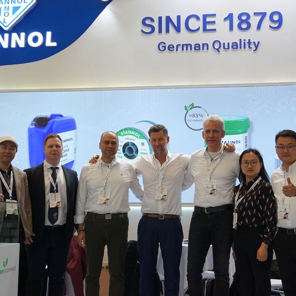 The picture shows a group of seven people in front of the Stannol stand. They are wearing formal clothes, have their arms around their shoulders and are smiling friendly into the camera.