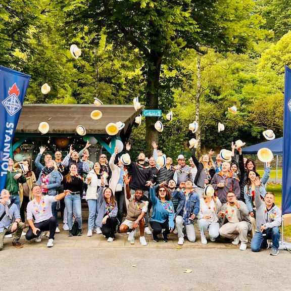 Auf dem Bild ist eine größere Gruppe von Menschen zu sehen, die Strohhüte in die Luft werfen. Im Hintergrund sieht man Bäume und eine Grillhütte.