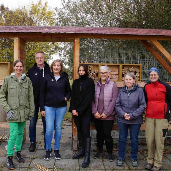 The picture shows a group of seven men and women in front of a roofed insect hotel.