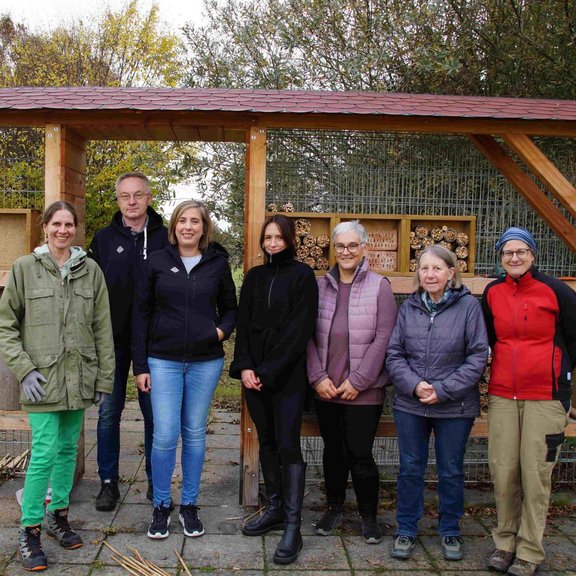 Auf dem Bild ist eine Gruppe von sieben Männern und Frauen vor einem überdachten Insektenhotel zu sehen.