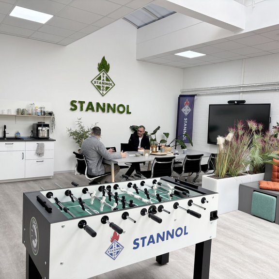 Auf dem Bild sieht man einen großen Schulungsraum Im Vordergrund steht ein Tischkicker, rechts sind Sitzreihen mit orangenen Kissen zu sehen. Im Hintergrund steht ein Tisch, an dem zwei Personen sitzen. Rechts hinten ist eine weiße Küchenzeile und auf der linken Seite ein großer Monitor zu sehen. An der WAnd im Hintergrund hängt ein großes Stannol-Logo aus Moos. 