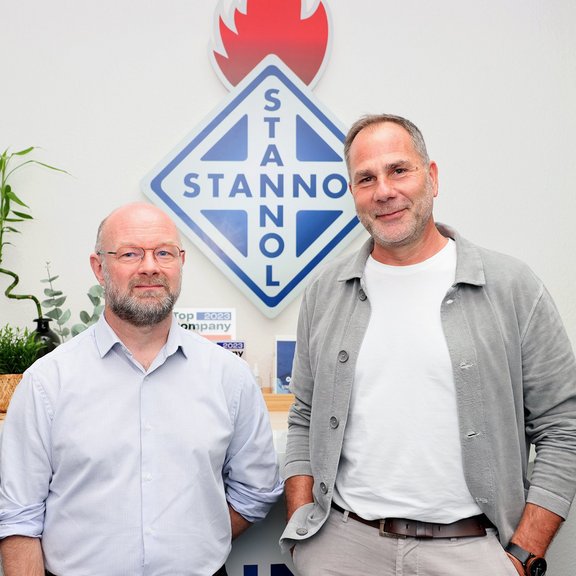 The picture shows two men in front of a large Stannol logo: Jean-Emmanuel Lechene is on the left, Thomas Kolossa is on the right. Both are smiling friendly into the camera.