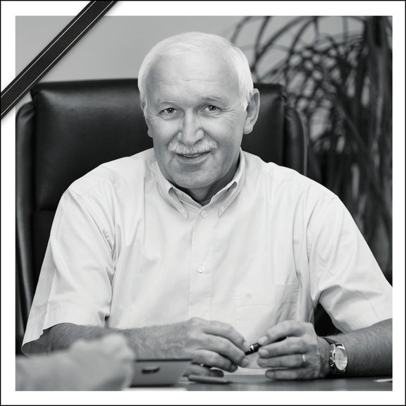 The black and white picture shows an older gentleman with white hair and a white shirt sitting at a desk. He smiles friendly into the camera. The photo bears a mourning ribbon.