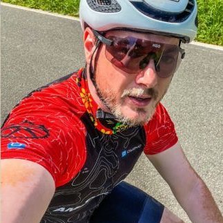 The picture shows a young man with a white cycling helmet, red and black jersey and cycling goggles on a racing bike. He is taking a selfie and smiling friendly into the camera.