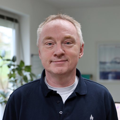 The picture shows a man with thin, grey hair. He is wearing a dark blue shirt with a white T-shirt underneath. He smiles friendly at the camera.
