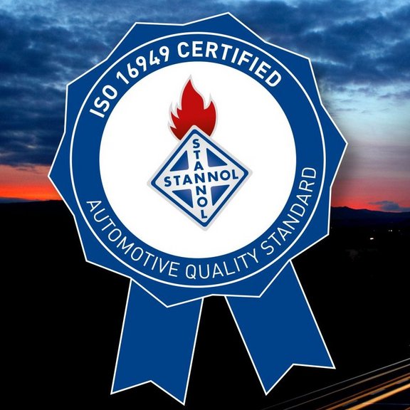 The image shows a badge-shaped logo representing a certification. The outer ring is blue with white text that reads "ISO 16949 Certified" at the top and "Automotive Quality Standard" at the bottom. Inside the circle is the logo of "Stannol." Below the circle is a blue ribbon with two ends, emphasizing the award-like character of the design. The background shows a sunset scene with a gradient of warm and dark tones.