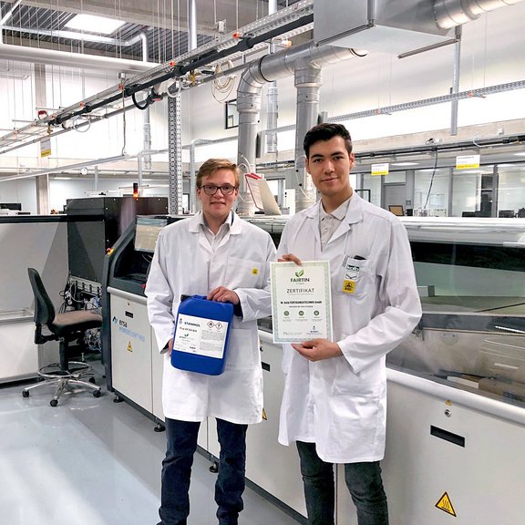 The picture shows two young men in a production hall. They are both wearing white coats. The man on the left is blonde and is holding a blue flux canister in his hands. The man on the right has dark hair and is holding a certificate up to the camera.