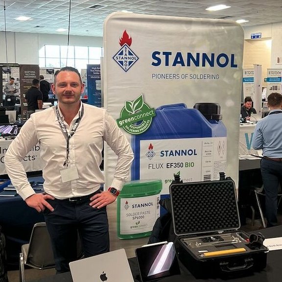 The picture shows a young man in a white shirt standing behind a small exhibition stand. To his right is a Stannol display with various products. 
