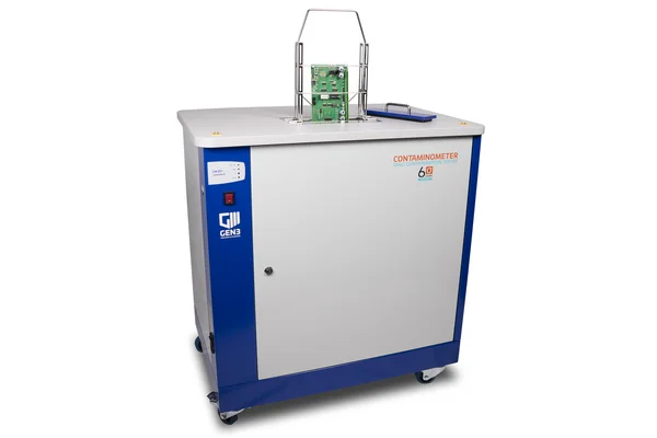 The picture shows a contaminometer device from Gen3 Systems: A large grey box with a holder on the top where circuit boards etc. can be lowered into the device.