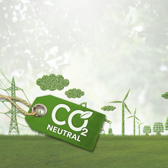 The image shows a symbolic representation of sustainability and climate neutrality. In the foreground, there is a green tag with the inscription "CO₂ Neutral." The tag is attached with a string and includes a leaf symbol within the lettering, emphasizing eco-friendly and climate-friendly aspects.  In the background, a green landscape stretches out with various elements made of plants and trees. These include wind turbines, power poles, and buildings, all crafted from foliage or grass. Additionally, there are clouds made of leafy structures. The background is slightly blurred, creating a peaceful, nature-oriented atmosphere. The image symbolizes a focus on renewable energy and CO₂ neutrality.