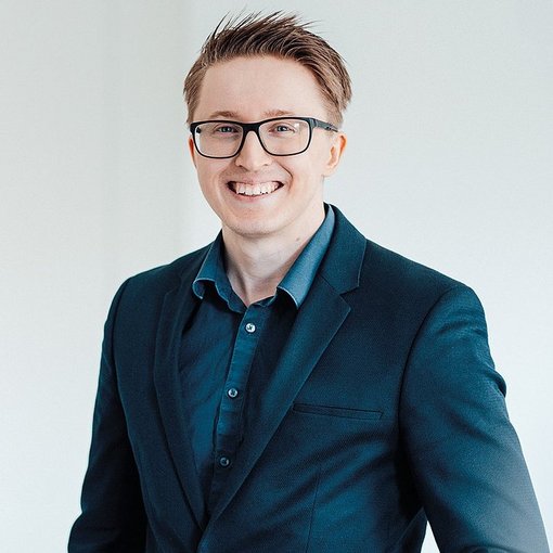 The picture shows a blond young man in a blue shirt and suit jacket. He is wearing glasses and is smiling friendly into the camera.
