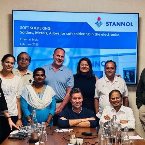 The picture shows a group of people in front of a screen with a PowerPoint presentation on Stannol. Marco Dörr, Thomas Kolossa and Batcheva Dominguez-Tellez are standing or sitting in the centre. They are surrounded by their Indian hosts. All of them are smiling friendly into the camera. 