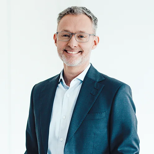 Auf dem Bild ist ein Mann mit hellem Hemd und blauem Sakko zu sehen. Er hat kurze graue Haare und trägt eine Brille. Er lächelt freundlich in die Kamera.