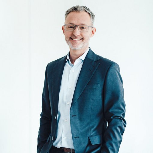 The picture shows a man wearing a light-coloured shirt and a blue suit jacket. He has short grey hair and wears glasses. He smiles friendly into the camera.