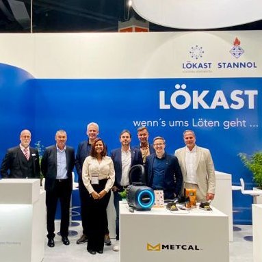 The picture shows a group of people in formal attire at a trade fair stand, smiling friendly into the camera. The Stannol and Lökast logos can be seen in the background.