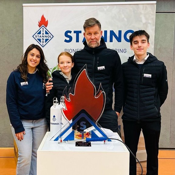 Auf dem Bild sind vier Personen vor einem Stannol-Banner zu sehen, die freundlich in die Kamera lächeln. Vor ihnen steht ein Tisch mit einem "heißen Draht" in Form der Stannol-Flamme. 