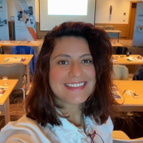 The picture shows a young woman with brown, shoulder-length hair in a white blouse. She is taking a selfie with a conference room in the background.