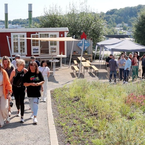 The picture shows a garden with a path on which several people are walking. A red construction trailer can be seen in the background. 