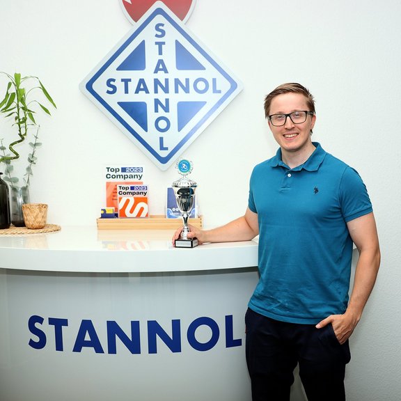 The picture shows a young blonde man with glasses at a Stannol counter. He is wearing a turquoise polo shirt, dark trousers and is holding a trophy in his hand.