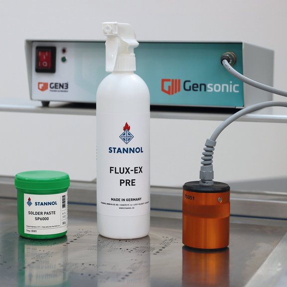 The picture shows the ultrasonic cleaning device Gensonic from Gen3 including an orange-coloured ultrasonic transducer, a green solder paste can with SP6000 solder paste from Stannol and a white spray bottle of Flux-Ex pre-cleaner from Stannol.