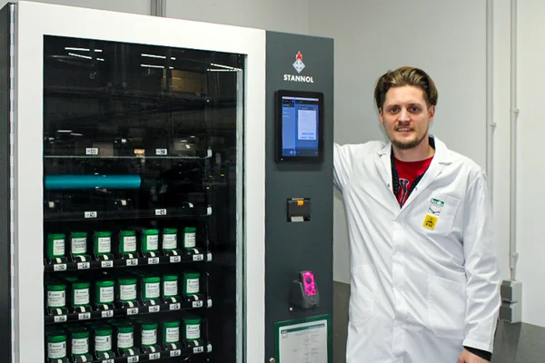 Auf dem Bild ist ein junger Mann neben dem Smart Warehouse zu sehen. Er trägt ein rotes Shirt und einen weißen Kittel. Das Smart Warehouse hat eine Glasfront, durch die man die verschiedenen Fächer mit Lotpasten-Dosen sehen kann. Rechts daneben befindet sich ein Eingabe-Display.