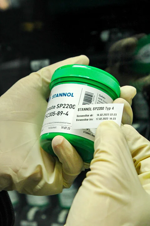 Auf dem Bild sind zwei Hände in Einmalhandschuhen zu sehen. Ein Aufkleber mit verschiedenen Daten wird auf eine grüne Lotpastendose geklebt.