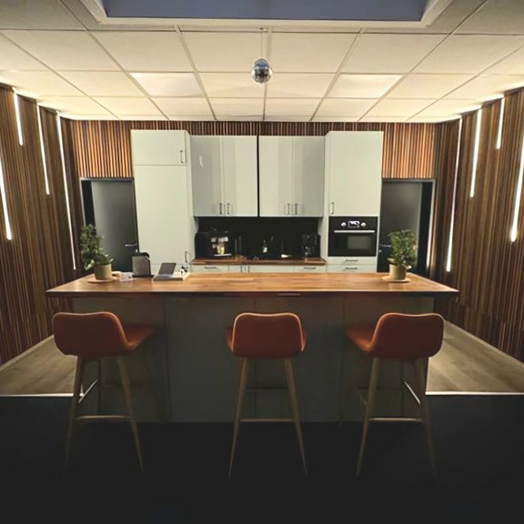 The picture shows a modern, light grey kitchen unit with a counter in the foreground and three orange bar stools.