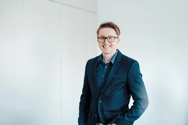 The picture shows a young blond man with glasses. He is wearing a dark blue suit and a blue shirt. He smiles friendly into the camera.