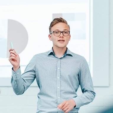 The picture shows a young blond man with glasses in a light-coloured shirt explaining something to an audience.