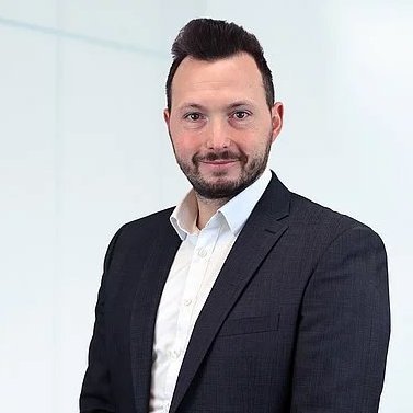 The picture shows a man in a white shirt and an anthracite-coloured jacket. He has dark hair and a beard. He smiles friendly into the camera.