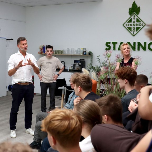 Eine Gruppe junger Menschen sitzt aufmerksam in einem modernen, hellen Raum bei Stannol. Ein Mann im weißen Hemd spricht engagiert vor der Gruppe, während zwei weitere Erwachsene im Hintergrund stehen und zuhören. Auf der Wand ist das Stannol-Logo zu sehen, daneben befinden sich Pflanzen und eine kleine Kaffeebar. 