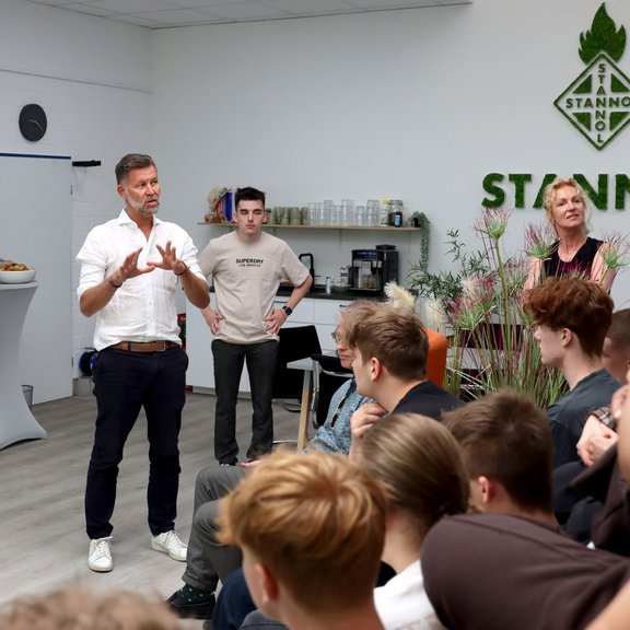 Eine Gruppe junger Menschen sitzt aufmerksam in einem modernen, hellen Raum bei Stannol. Ein Mann im weißen Hemd spricht engagiert vor der Gruppe, während zwei weitere Erwachsene im Hintergrund stehen und zuhören. Auf der Wand ist das Stannol-Logo zu sehen, daneben befinden sich Pflanzen und eine kleine Kaffeebar. 