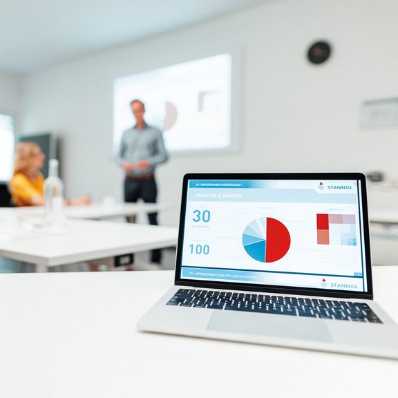 Auf dem Bild ist ein Seminarraum zu sehen, im Vordergrund steht ein aufgeklappter Laptop, im Hintergrund sitzen zwei Seminar-Teilnehmerinnen und der Seminarleiter erklärt etwas vor einer Leinwand.