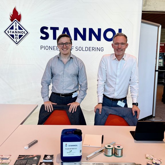 The picture shows two men behind a table smiling at the camera. Stannol products such as a canister of flux, a solder bar and spools of solder wire can be seen on the table. The Stannol logo can be seen in the background. 