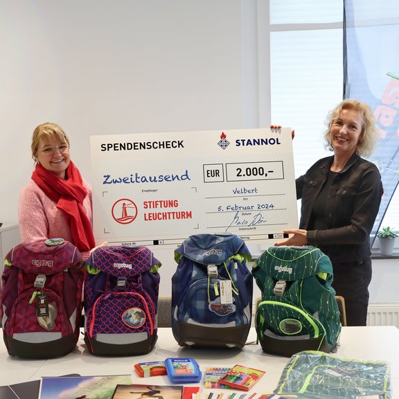 The picture shows two women standing behind a table and holding up a cheque together. On the table are four schoolbags and other school materials.