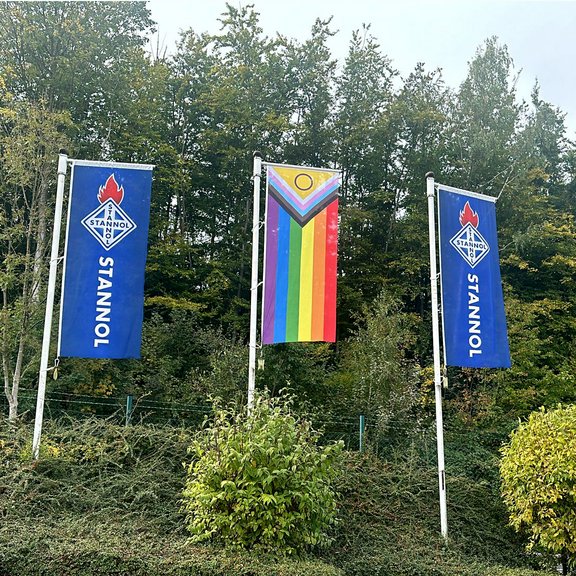 Auf dem Bild sind drei Flaggen zu sehen, rechts und links zwei blaue Stannol-Flaggen mit Logo und in der Mitte eine Regenbogenflagge.