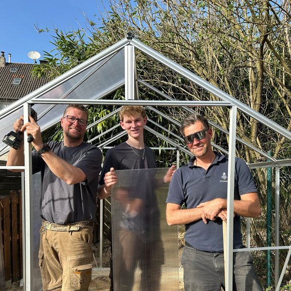 The picture shows three men in work clothes setting up a small greenhouse together. They are smiling friendly into the camera.