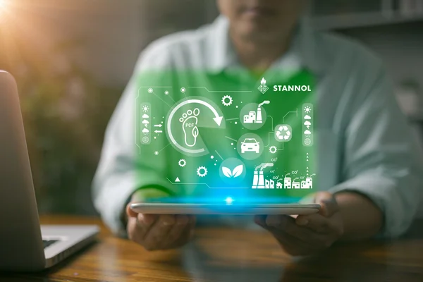 Das Bild zeigt symbolisch, dass die Firma Stannol den Product Carbon Footprint (PCF) für ihre Produkte ausweist. Eine Person hält ein Tablet, über dem ein holografisches, grünes Display mit dem Stannol-Logo in der oberen rechten Ecke schwebt. Das Display zeigt Symbole für Nachhaltigkeit und Umweltschutz, wie einen Fußabdruck, ein Blatt, ein Recycling-Symbol und CO₂-Reduktion. Die unscharfe Darstellung der Person im Hintergrund lenkt den Fokus auf die Nachhaltigkeitssymbole und das Engagement von Stannol für transparente Umweltinformationen.