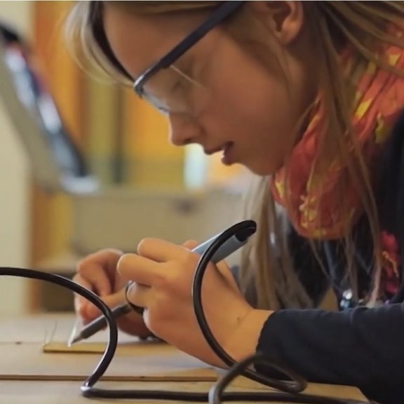 Auf dem Bild ist ein Mädchen mit braunen Haaren und einer Scutzbrille zu sehen, die einen Lötkolben in der Hand hält. Sie schaut konzentriert auf ihre Arbeit.